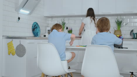 Mom-in-the-kitchen-washing-dishes-and-two-sons-sitting-at-a-Desk-drawing-on-paper-with-colored-pencils.